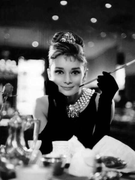 Portrait culte en noir et blanc d'Audrey Hepburn (Holly Golightly) accoudée à une table, portant un collier de perles, un diadème et tenant son long porte-cigarette.