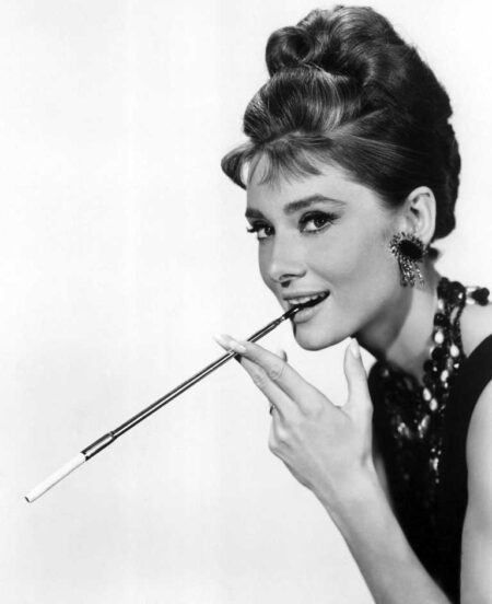 Portrait noir et blanc iconique d'Audrey Hepburn dans Diamants sur canapé, posant avec élégance et tenant un long porte-cigarette.