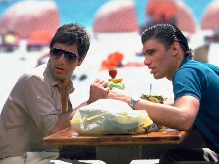 Al Pacino (Tony Montana) et Steven Bauer (Manny) assis à une table de plage à Miami avec des cocktails, scène du film Scarface, ambiance années 80 colorée.
