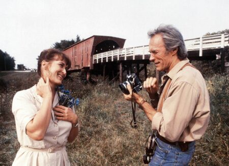 Photo couleur de Meryl Streep et Clint Eastwood dans le film Sur la route de Madison. Elle sourit avec des fleurs bleues à la main tandis qu'il s'apprête à la photographier devant un pont couvert en bois.