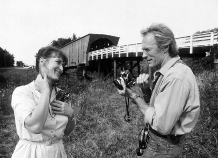 Photographie noir et blanc de Clint Eastwood tenant un appareil photo face à Meryl Streep souriante, devant un pont couvert dans Sur la route de Madison.