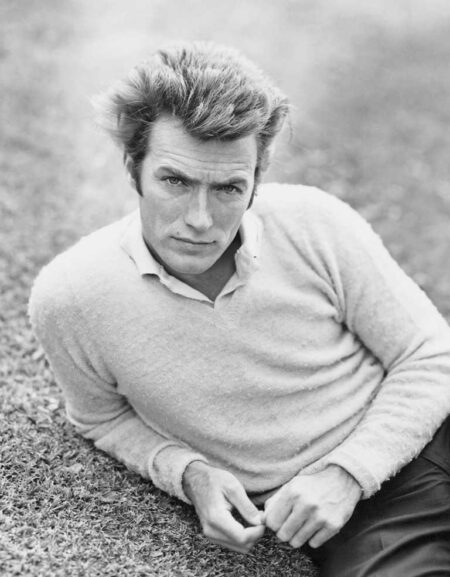 Portrait en noir et blanc d'un jeune Clint Eastwood allongé sur l'herbe, portant un pull clair sur une chemise à col, fixant l'objectif avec intensité.