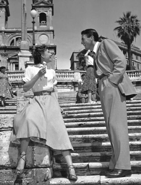 Photographie en noir et blanc d'Audrey Hepburn mangeant une glace sur les marches de la Place d'Espagne à Rome, face à Gregory Peck qui lui sourit.