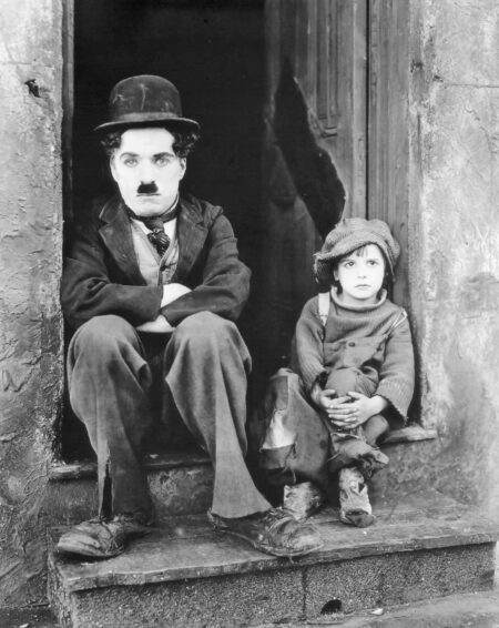 Photographie iconique en noir et blanc de Charlie Chaplin et du jeune acteur Jackie Coogan assis côte à côte sur un perron, dans leurs costumes fidèles au film Le Kid.