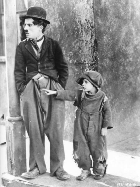 Photographie de plateau en noir et blanc de Charlie Chaplin (Charlot) s'appuyant contre un poteau, tandis que son jeune partenaire pointe du doigt vers la gauche, tous deux vêtus de leurs costumes fidèles au film Le Kid.