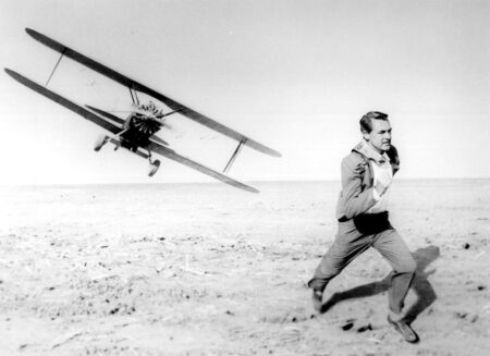 Photographie de plateau en noir et blanc du film La Mort aux trousses (1959) montrant Cary Grant en costume, courant dans un champ désertique alors qu'un avion biplan le poursuit à basse altitude en arrière-plan.