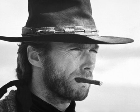 Portrait serré en noir et blanc de Clint Eastwood en cowboy, portant un chapeau et un cigare aux lèvres, le regard plissé vers l'horizon.