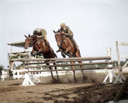 Photographie historique colorisée de 1925 montrant deux cavaliers et leurs chevaux franchissant simultanément un obstacle en bois lors d'un concours hippique.