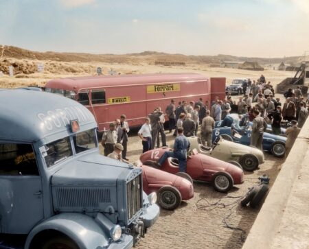 Photographie historique colorisée en vue plongée depuis une tribune, montrant quatre monoplaces de course de 1952 (dont une Ferrari rouge à l'arrière, une bleue, une grise et une verte) préparées par leurs mécaniciens dans la voie des stands, entourées d'une foule en costume.