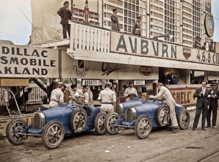 Photographie historique colorisée du Grand Prix d'Espagne 1926, montrant des mécaniciens préparant deux légendaires Bugatti bleues dans la voie des stands, sous des panneaux publicitaires d'époque.