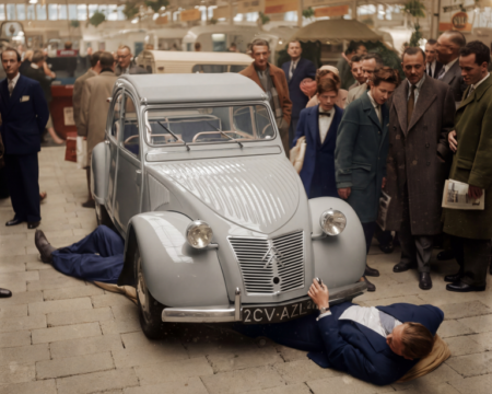 Photographie historique colorisée de 1958 montrant des hommes en costume allongés sur le sol pour inspecter le dessous d'une Citroën 2CV grise lors d'un salon automobile, entourés d'une foule curieuse.