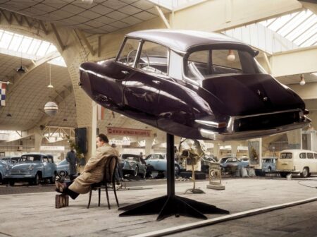 Photographie historique colorisée d'un salon automobile en 1958 montrant la carrosserie d'une Citroën DS aubergine exposée en lévitation sur un pied, avec un homme assis calmement en dessous.