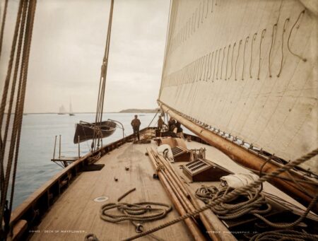 Photographie historique colorisée en vue subjective depuis le pont en bois du voilier Mayflower en 1886, montrant les cordages, l'immense voile et l'équipage.