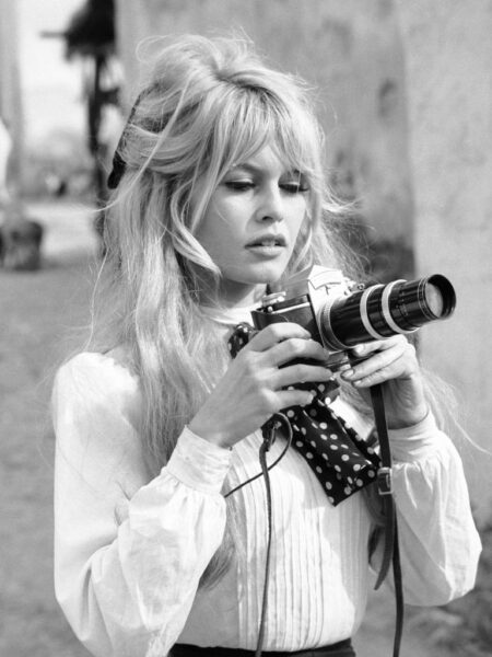 Photographie en noir et blanc de Brigitte Bardot portant une blouse blanche et un foulard à pois, tenant un appareil photo professionnel avec un long téléobjectif.