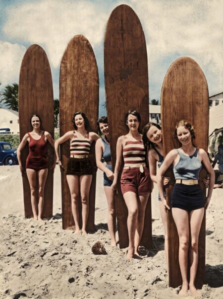Photographie historique colorisée montrant un groupe de six jeunes femmes souriantes en maillots de bain rétro, posant sur la plage avec d'immenses planches de surf en bois massif.