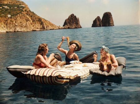 Photographie vintage colorisée montrant trois femmes riant et mangeant des spaghettis sur des matelas gonflables flottant sur la mer à Capri.