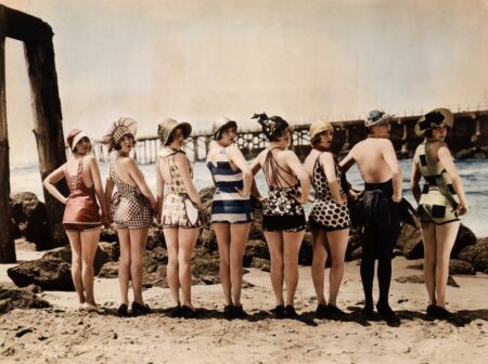 Photographie vintage colorisée de 1920 montrant huit femmes en maillots de bain rétro alignées sur la plage et regardant par-dessus leur épaule.