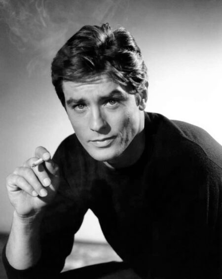Portrait photographique vintage en noir et blanc de l'acteur français Alain Delon jeune, fixant l'objectif avec un regard intense et tenant une cigarette allumée.
