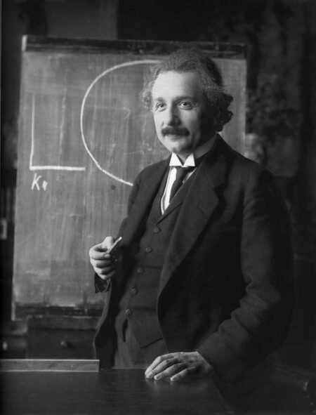 Photographie en noir et blanc d'Albert Einstein debout devant un tableau noir, tenant une craie à la main.