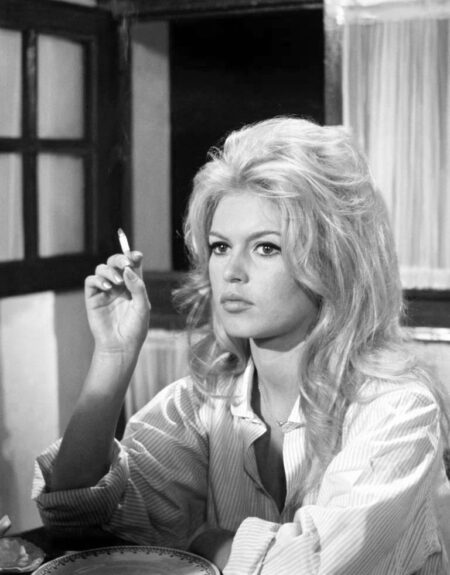 Portrait en noir et blanc de Brigitte Bardot en 1962, portant une chemise rayée et tenant une cigarette, avec un regard mélancolique et sa coiffure blonde signature.