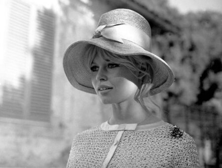 Portrait lumineux en noir et blanc de Brigitte Bardot en 1961, portant un grand chapeau clair avec un ruban, une tenue texturée et une broche, le visage partiellement ombragé.