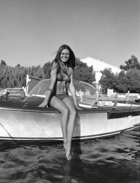 Photographie artistique en noir et blanc de Brigitte Bardot souriante, assise sur le bord d'un bateau devant sa villa La Madrague à Saint-Tropez en 1973.