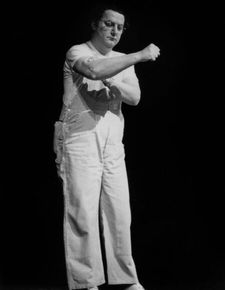 Photographie en pied et en noir et blanc de Coluche sur scène en 1977, portant sa célèbre salopette à rayures, un t-shirt blanc et des lunettes rondes, effectuant un geste humoristique sur un fond noir profond.