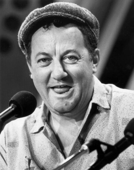 Photographie en noir et blanc de l'humoriste français Coluche en 1985, souriant face à des micros et portant sa célèbre casquette gavroche en tweed.