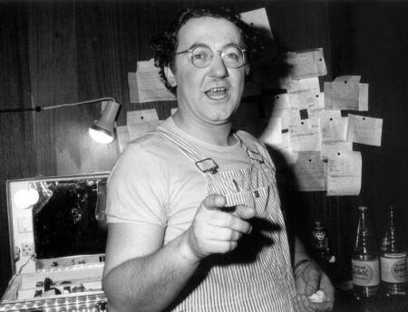 Photo noir et blanc de Coluche en 1975, portant des lunettes rondes et une salopette rayée, pointant du doigt vers l'appareil photo depuis sa loge avec des feuillets épinglés au mur et une mallette de maquillage éclairée.