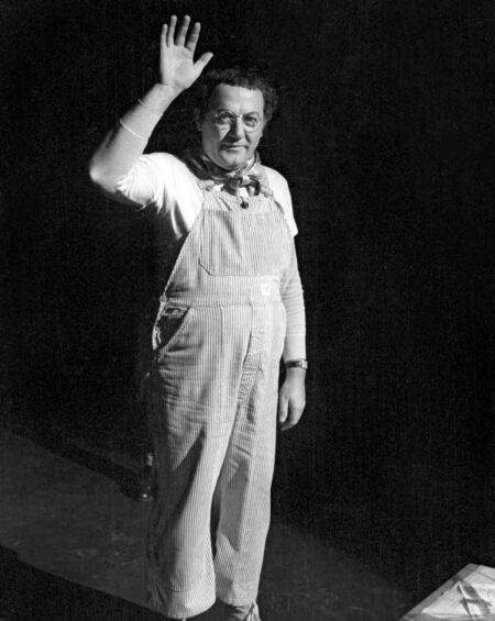 Photographie en noir et blanc de Coluche sur scène en 1981, vêtu de sa salopette à rayures et d'un foulard, levant la main pour saluer le public avec une table visible au premier plan.