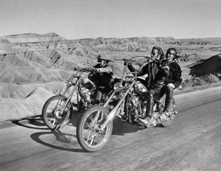 Photographie en noir et blanc du film Easy Rider (1969) montrant Dennis Hopper, Peter Fonda et Jack Nicholson sur des motos choppers. Ils roulent sur une route dégagée au milieu d'un paysage de canyons désertiques. Jack Nicholson est assis à l'arrière de la moto de Peter Fonda.