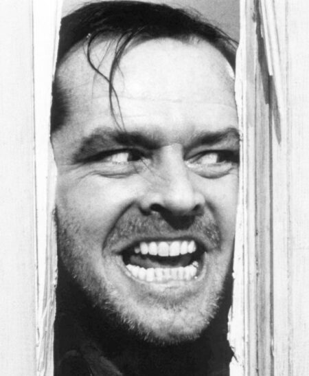 Gros plan en noir et blanc de Jack Nicholson (Jack Torrance) dans le film The Shining. Il passe son visage à travers la fente d'une porte en bois fracassée, affichant un sourire dément et un regard maniaque.