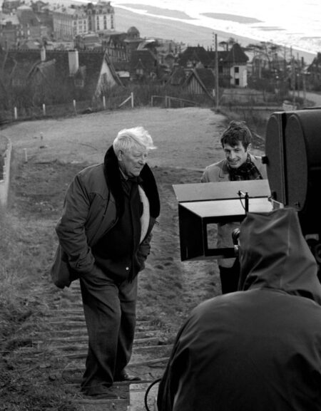 Photographie en noir et blanc sur le tournage du film Un Singe en Hiver, montrant Jean Gabin et Jean-Paul Belmondo debout sur des marches en extérieur avec un village côtier en arrière-plan, face à une caméra de cinéma et un membre de l'équipe.