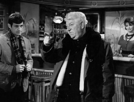 Photographie en noir et blanc du film Un Singe en Hiver avec Jean Gabin en manteau épais pointant le doigt, observé par Jean-Paul Belmondo dans un décor de bar avec une barmaid en arrière-plan.