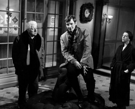 Affiche noir et blanc du film Un Singe en Hiver montrant Jean Gabin de face, observant Jean-Paul Belmondo qui est assis au-dessus de lui sur le dossier d'un gros pouf en cuir capitonné, dans un grand hall avec des portes vitrées. Une femme en robe de chambre sombre les regarde depuis l'arrière-plan à droite.