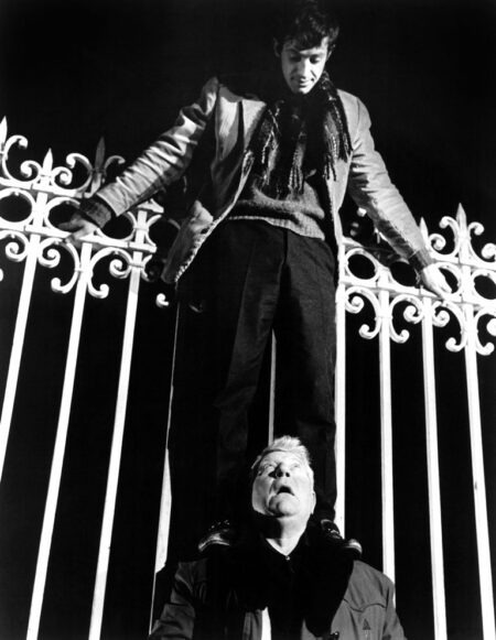 Photographie en noir et blanc du film Un Singe en Hiver montrant le jeune Jean-Paul Belmondo debout sur les épaules de Jean Gabin pour tenter d'escalader une haute grille en fer forgé pendant la nuit.