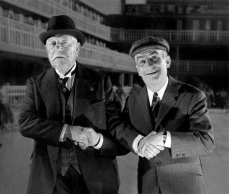 Photographie en noir et blanc de Jean Gabin en costume et chapeau serrant la main de Louis de Funès souriant en casquette, issue du film Le Tatoué.