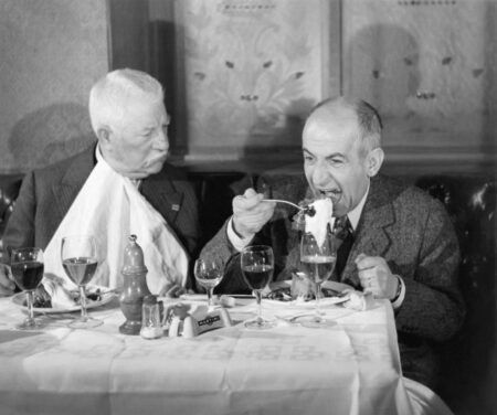 Photographie en couleur du film Le Tatoué montrant Jean Gabin, l'air impassible avec une serviette nouée au cou, observant Louis de Funès qui mange goulûment à une table de restaurant dressée avec des verres de vin.