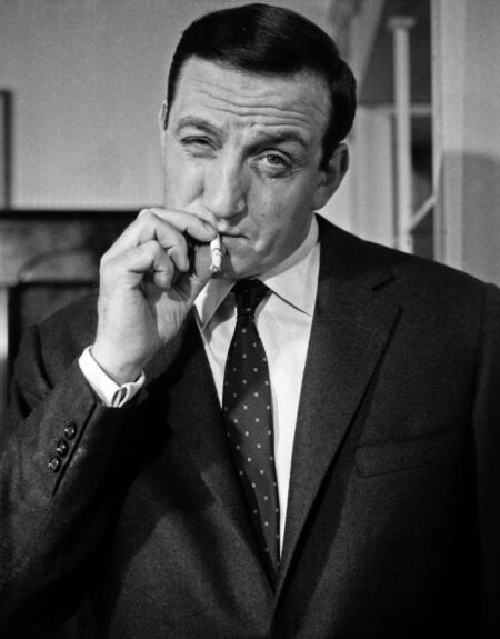 Portrait en noir et blanc de Lino Ventura dans le rôle de Fernand Naudin. Il est de face, vêtu d'un costume sombre et d'une cravate, avec une expression sérieuse et déterminée.