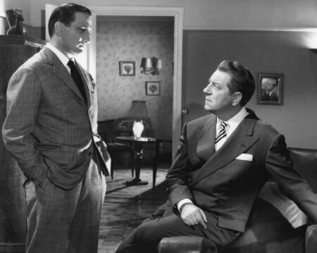 Photographie noir et blanc du film Touchez pas au grisbi : Jean Gabin assis sur un canapé regarde Lino Ventura debout, tous deux en costume élégant dans un décor de salon d'époque.