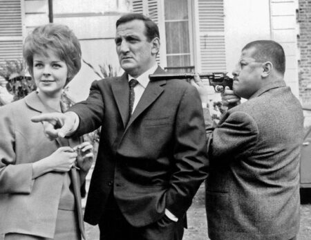 Photographie de plateau en noir et blanc des Tontons Flingueurs : Lino Ventura pointe du doigt aux côtés de Sabine Sinjen, tandis que Francis Blanche vise avec un revolver à silencieux devant une maison.
