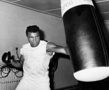 Affiche Mohamed Ali : photographie d'action en noir et blanc de 1963 montrant Mohamed Ali (Cassius Clay) portant un coup puissant dans un sac de frappe historique "Ampro London" en cuir.