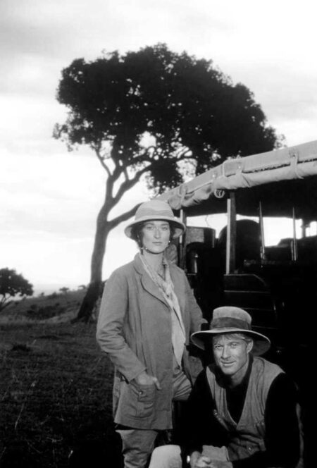 Photographie de plateau en noir et blanc du film Out of Africa montrant Meryl Streep et Robert Redford en tenue de safari. Meryl Streep est debout, les mains dans les poches de sa veste, tandis que Robert Redford est accroupi à ses côtés. Ils posent devant un véhicule de brousse et un grand acacia dans un paysage de plaine africaine.