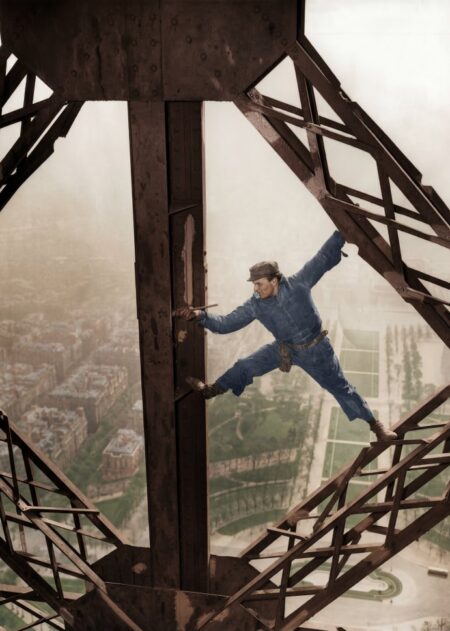Photographie historique colorisée de 1953 montrant un peintre en bâtiment en équilibre sur la structure de la Tour Eiffel, un pinceau à la main, surplombant Paris.