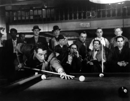 Photographie de plateau en noir et blanc du film L'Arnaqueur (1961) montrant Paul Newman penché sur une table de billard, une cigarette aux lèvres, s'apprêtant à tirer sous le regard d'un groupe d'hommes dans une salle de jeu sombre.