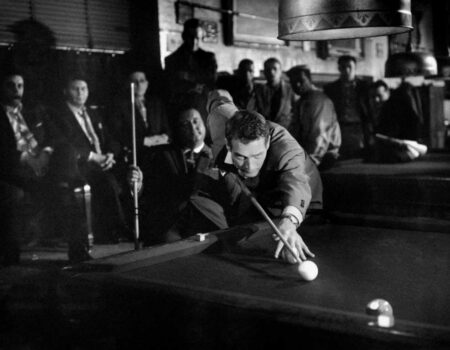 Photo noir et blanc du film L'Arnaqueur (1961) montrant Paul Newman en pleine action, le corps très incliné au-dessus d'une table de billard pour préparer un coup de queue. Un groupe d'hommes l'observe avec attention dans l'ombre d'une salle de jeu enfumée.