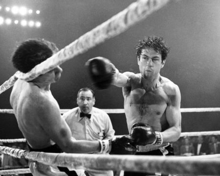 Photographie en noir et blanc du film Raging Bull montrant Robert De Niro (Jake LaMotta) sur un ring de boxe, en plein effort, assénant un coup de poing puissant à son adversaire sous le regard attentif de l'arbitre. La scène est marquée par un éclairage dramatique et des projections de sueur capturées en plein mouvement.