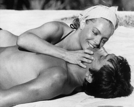 Photographie de cinéma en noir et blanc montrant Alain Delon et Romy Schneider allongés l'un contre l'autre dans une scène intime du film La Piscine.