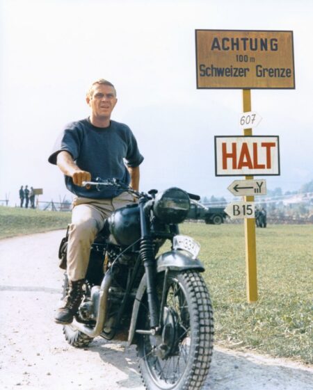 Photographie en couleur de l'acteur Steve McQueen vêtu d'un pull bleu, assis sur une moto vintage devant des panneaux jaunes de la frontière suisse, tirée du film La Grande Évasion.
