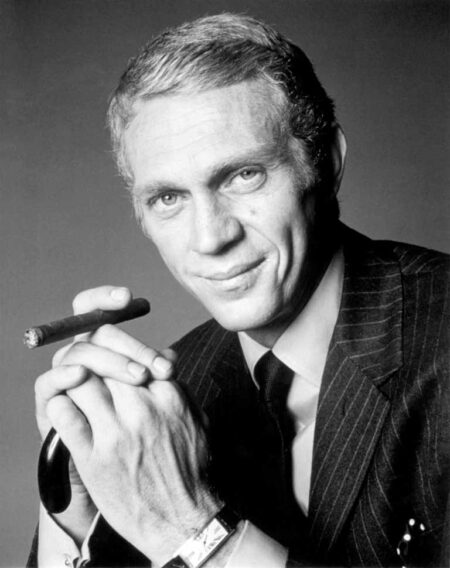 Portrait en noir et blanc de Steve McQueen dans le film L'Affaire Thomas Crown. L'acteur, très élégant en costume à rayures et cravate, regarde l'objectif avec un sourire en coin tout en tenant un cigare entre ses mains jointes.